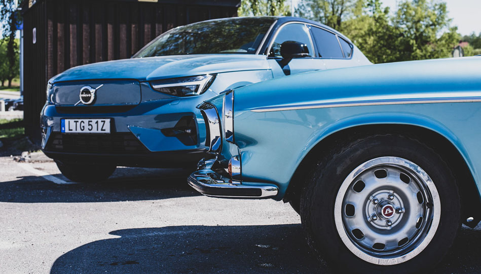 Två ljusblå bilar på en parkering. Volvo P1800 och en ny EC40 i bakgrunden.